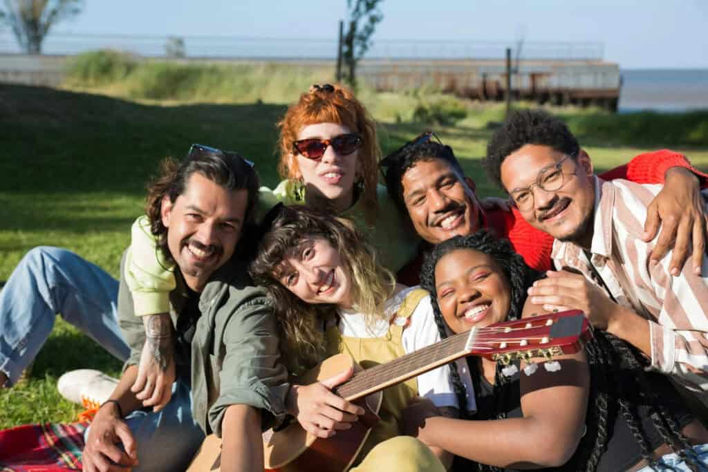 group of young adults sitting on blanket on a grassy field with a guitar and smiling 