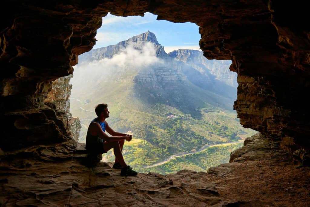 man sitting inside of a cave looking out into the sun with a mountain backdrop