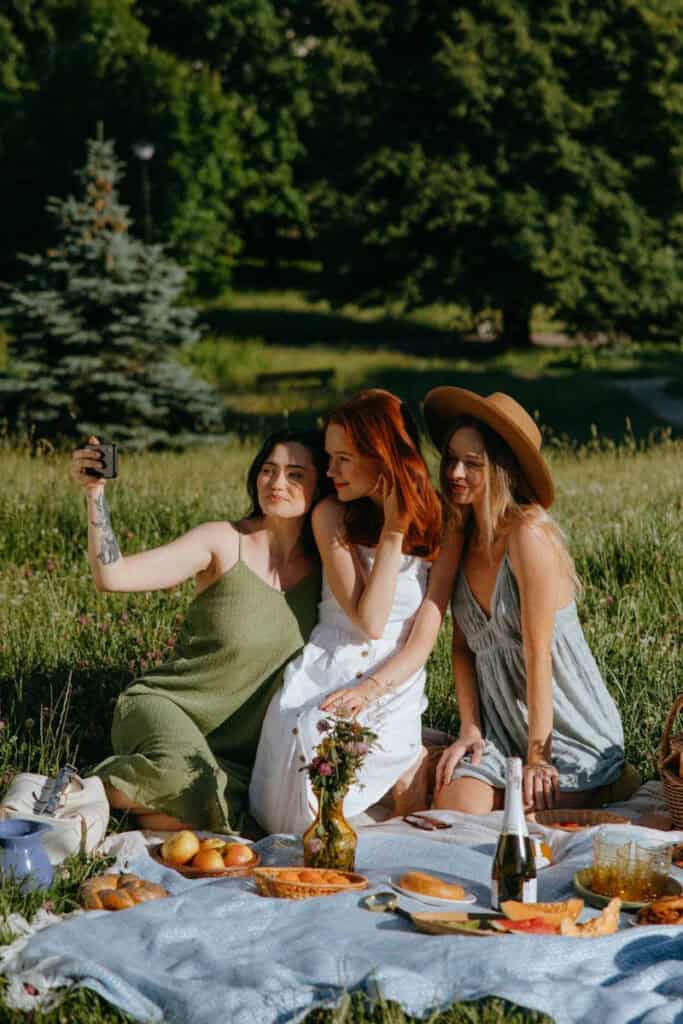 3 young women taking a selfie on a picnic blanket with food in front of them and trees / grass in background