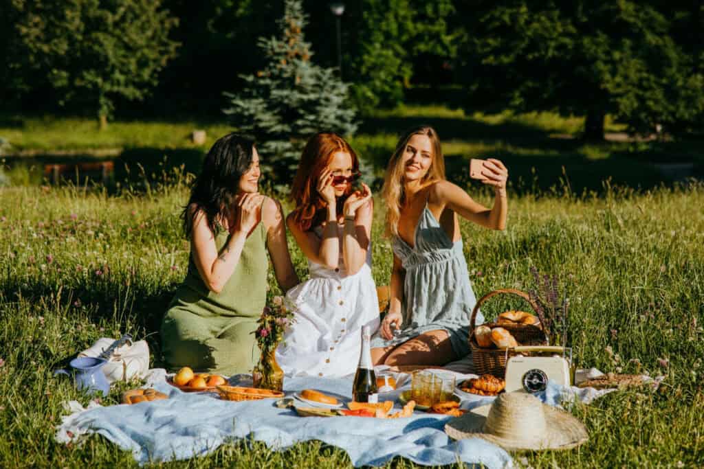 3 women taking a selfie on a picnic blanket with food in front and trees and grass in the background