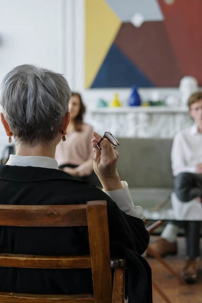 the back of a therapists head in the foreground with a man and woman sitting on a couch across from her during a therapy session 