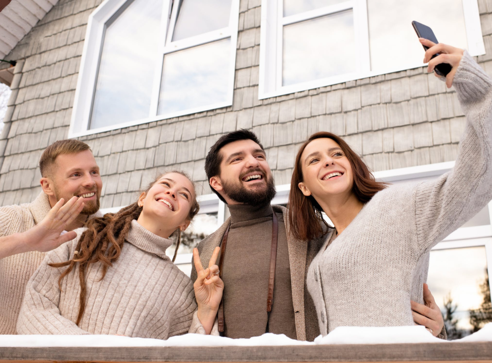 2 men and 2 women in tan shirts pose for a selfie in front of a snow covered railing on a porch in front of a house with tan shingles