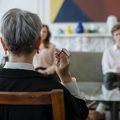 in the foreground you see the back of the head of an older adult therapist and she is looking at a couple sitting on a couch directly across from her in the background