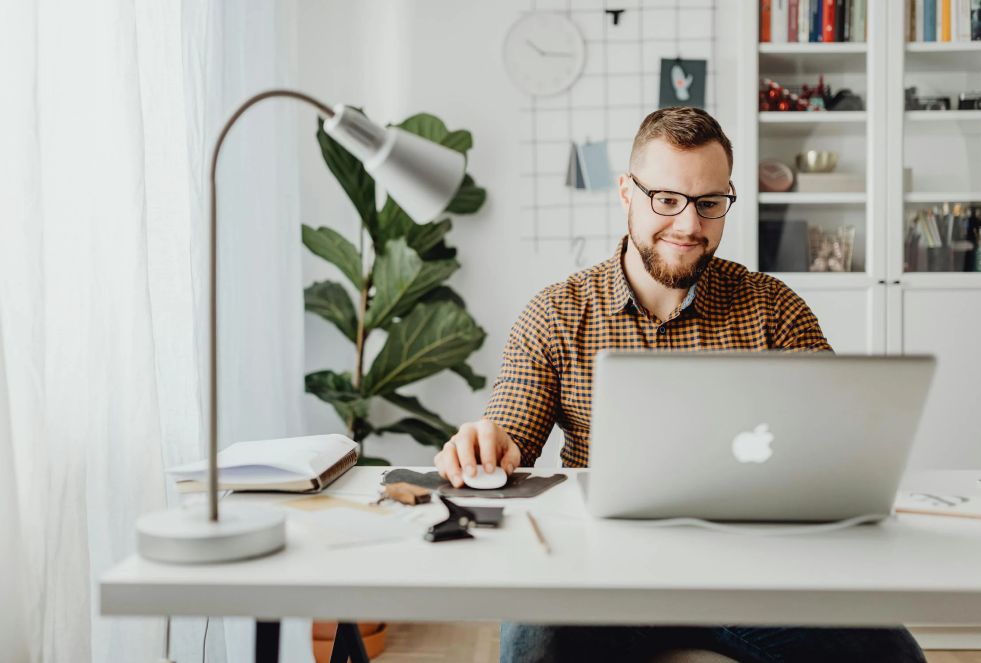 man sits at a white desk in a white office with book shelves behind him while looking at his laptop and smiling