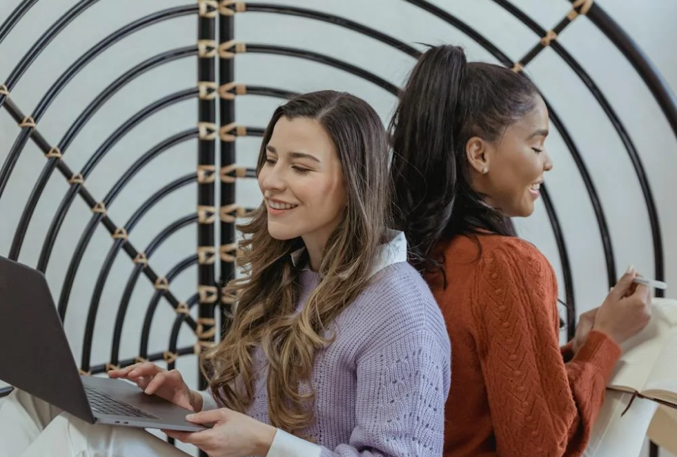 2 women are sitting back to back, one on a laptop and the other with a notepad with a geometrical room divider behind them