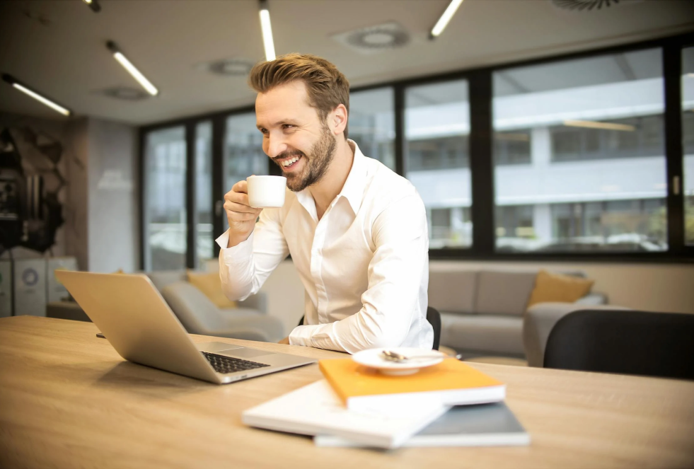 man sits at a desk in a high rise (city parking garage visible out the window) and is holding a coffee cup up to his face with his right hand while smiling at his laptop screen