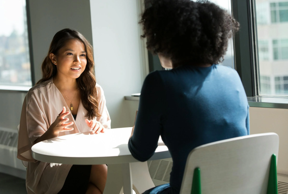 2 women sit across from each other at a table chatting in an office building high rise (city buildings visible out the window)