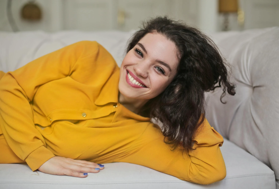 woman in a yellow blouse is laying on an ivory couch posing for a picture