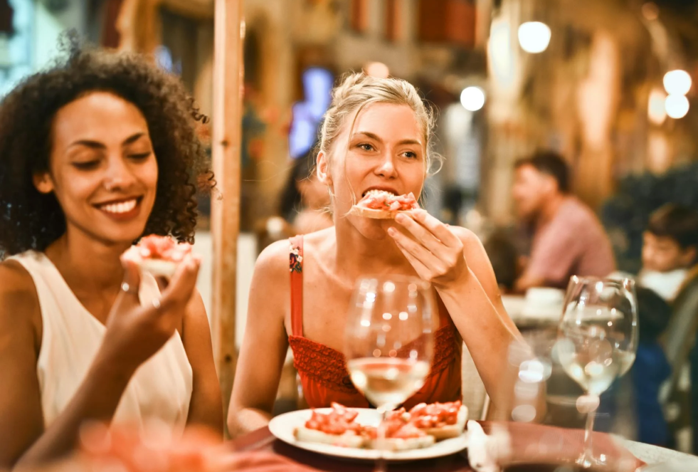 2 women sit next to each other in a restaurant sharing a plate of pizza and smiling with wine glasses in front