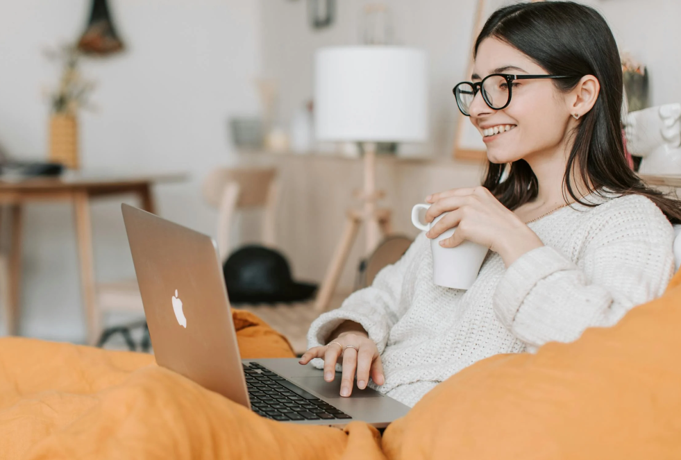 young adult woman with a white sweatshirt and glasses is sitting on her couch at home with a coffee mug in one hand and her other hand on her laptop trackpad, smiling at the screen