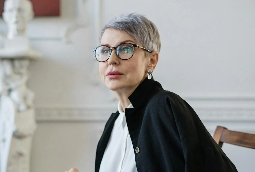 gray haired older woman therapist with a white blouse and black jacket and glasses peers into the distance - background is a white wall