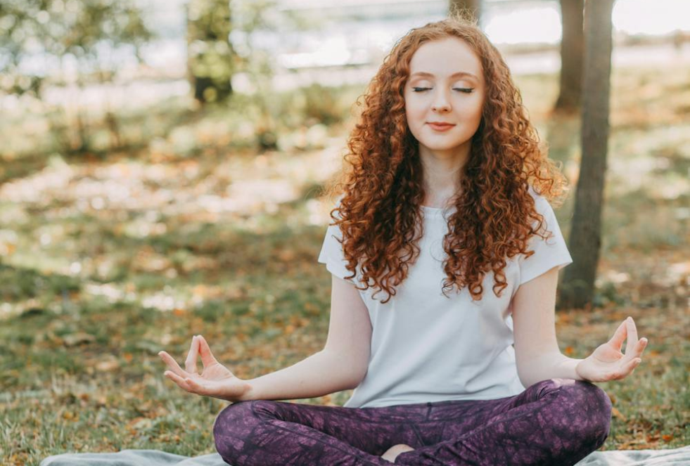 woman sitting in crosslegged yoga position with hands outstretched and palms facing up - thumbs touching index finger to form an o - background is a grassy area with trees