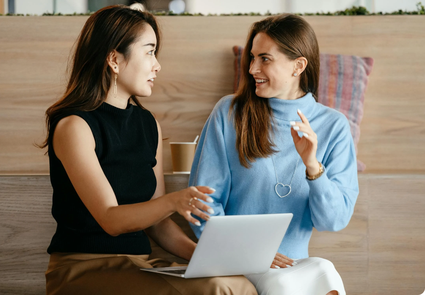 2 young women sitting next to each other on a wooden bench talking with a laptop in front of them