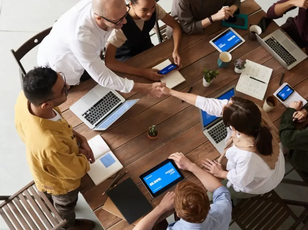 a group of 6 men and 2 women sitting on a table with their laptops and tablets. one woman and one man shaking hands.