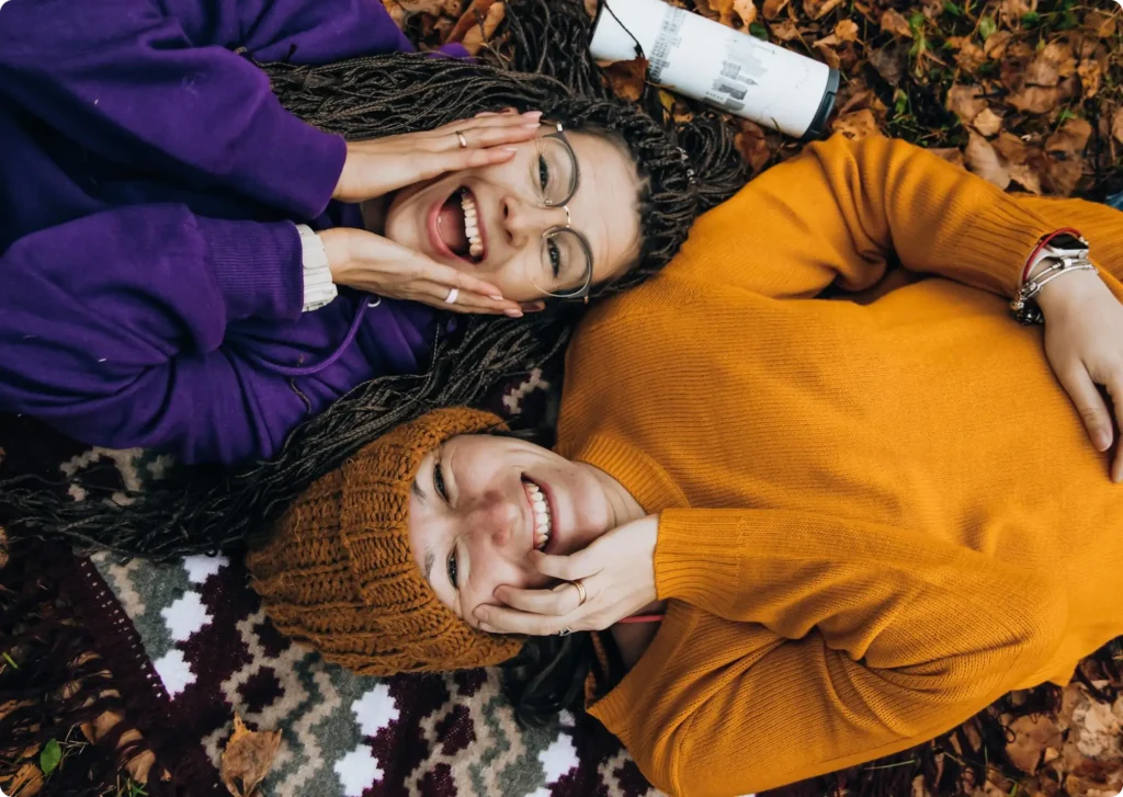 two ladies lying on a picnic blanket surrounded with fallen leaves smiling for the camera